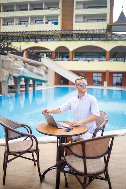 Hombre de negocios masculino guapo exitoso en gafas de sol trabaja en una computadora portátil sentado cerca de la piscina. Trabajo remoto. Persona de libre dedicación