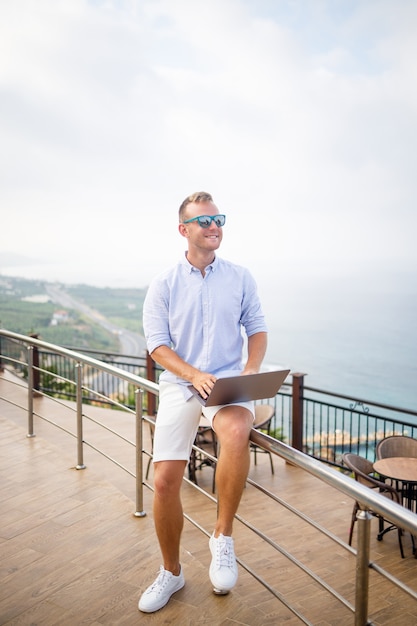 Hombre de negocios masculino joven acertado hermoso que trabaja con la computadora portátil mira el mar Mediterráneo. Lleva camisa y pantalones cortos blancos. Trabajo a distancia en vacaciones. Concepto de vacaciones