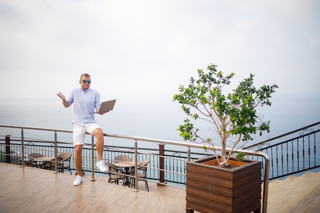 Hombre de negocios masculino joven acertado hermoso que trabaja con la computadora portátil mira el mar Mediterráneo. Lleva camisa y pantalones cortos blancos. Trabajo a distancia en vacaciones. Concepto de vacaciones