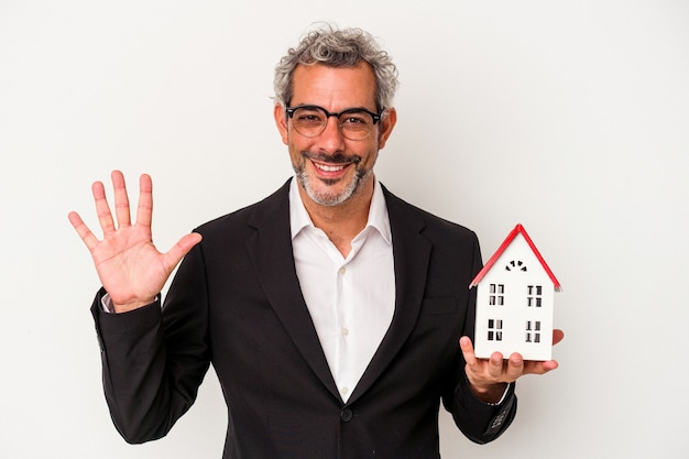 Hombre de negocios de mediana edad con facturas y modelo de casa aislado sobre fondo azul sonriendo alegre mostrando el número cinco con los dedos.