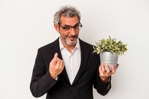 Hombre de negocios de mediana edad sosteniendo una planta aislada sobre fondo blanco apuntando con el dedo hacia usted como si estuviera invitando a acercarse.