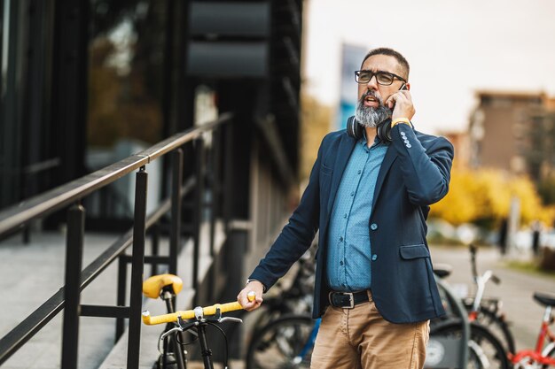 Un hombre de negocios de mediana edad va a trabajar en bicicleta y habla por teléfono inteligente frente al distrito de oficinas.