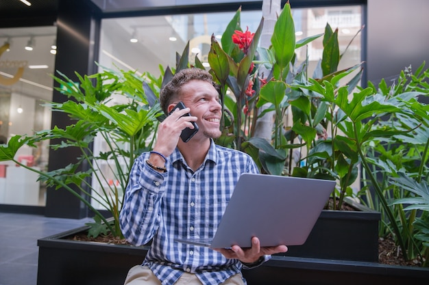 Hombre de negocios milenario habla por teléfono mientras sostiene su computadora portátil. Sonriente persona exitosa en el trabajo - elegante hombre caucásico durante una llamada telefónica.