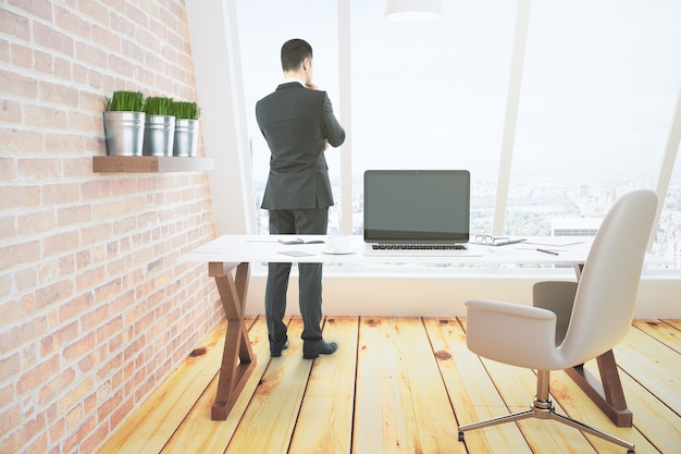 El hombre de negocios mira por la ventana en la habitación del loft con una computadora portátil con una pantalla en blanco simulada