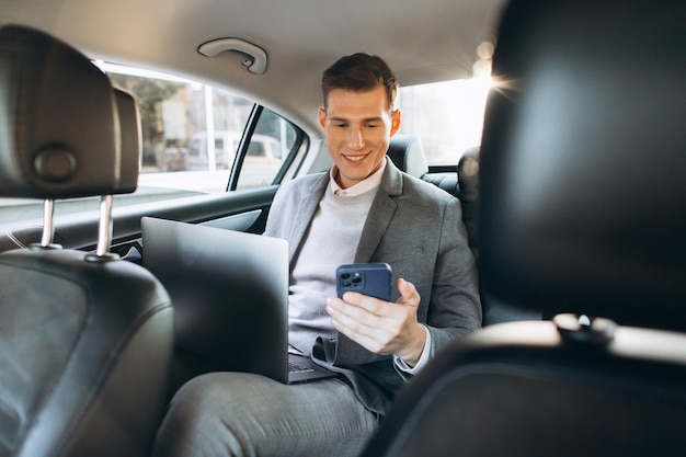 Un hombre de negocios moderno está hablando por un teléfono móvil y usando una tableta mientras está sentado en el asiento trasero de un auto