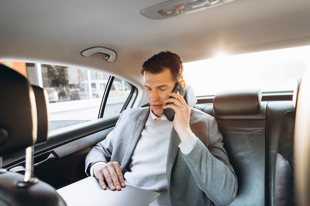 Hombre de negocios moderno hablando por un teléfono móvil y usando una computadora portátil mientras está sentado en el asiento trasero de un auto