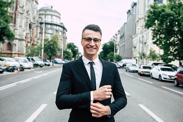 Hombre de negocios moderno Hombre joven confiado en traje completo ajustando su manga mientras está de pie al aire libre con el paisaje urbano en el fondo