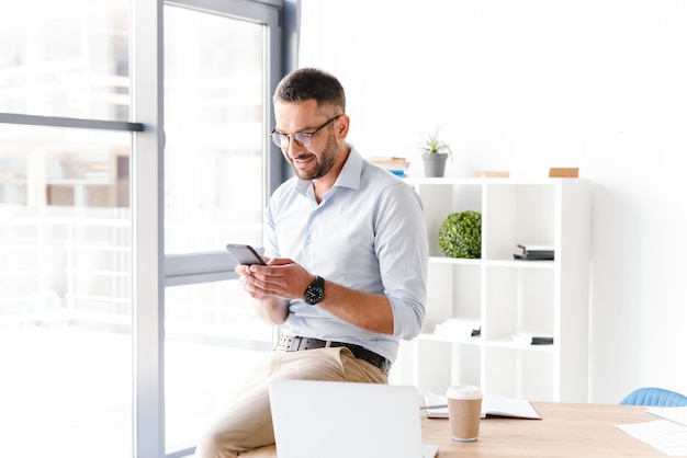 Foto hombre de negocios moderno en ropa formal sentado en la mesa en la oficina cerca de una ventana grande y usando el teléfono inteligente para el trabajo