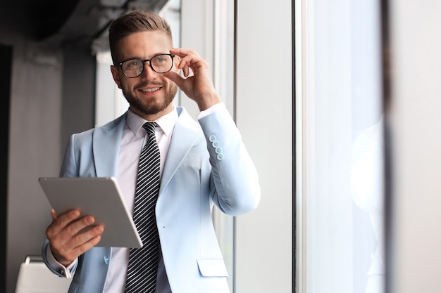 Foto hombre de negocios moderno en ropa formal con tableta digital mientras está de pie junto a la ventana en la oficina.