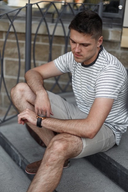 Hombre de negocios moderno con ropa de verano elegante se sienta en una escalera de piedra y mira el reloj.