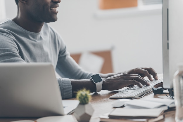 Hombre de negocios moderno en el trabajo. Primer plano de un joven africano guapo trabajando en la computadora y sonriendo mientras está sentado en su lugar de trabajo en la oficina en casa