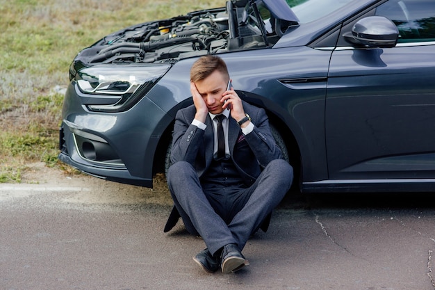 Hombre de negocios molesto usa teléfono sentado en la carretera cerca del coche roto