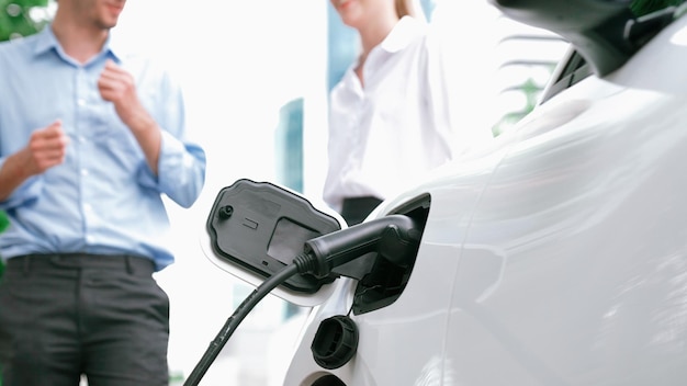 Un hombre de negocios y una mujer de negocios progresistas instalan el enchufe del cargador desde la estación de carga al coche eléctrico antes de conducir por el centro de la ciudad Coche recargable ecológico impulsado por energía limpia