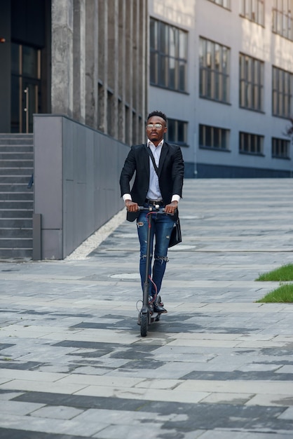 Hombre de negocios negro alegre moderno monta scooter eléctrico después de terminar su día de trabajo en la oficina.