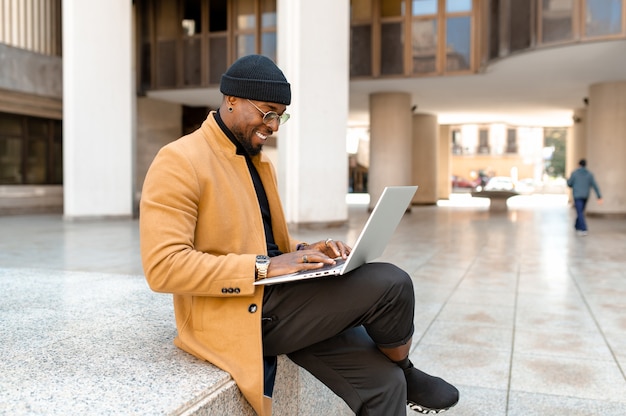 Hombre de negocios negro de retrato de cuerpo completo en ropa elegante y elegante sentado en pasos en la ciudad sonriendo y usando computadora portátil.