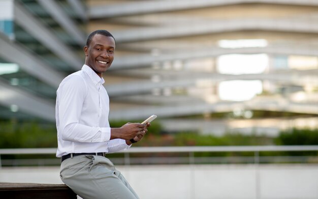 Un hombre de negocios negro sonriente con un teléfono inteligente en las manos parado al aire libre contra el centro de oficinas