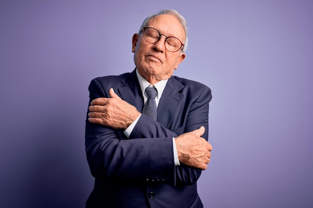 Hombre de negocios de pelo gris con gafas y traje elegante y corbata sobre fondo morado abrazándose feliz y positivo sonriendo confiado Amor propio y cuidado personal