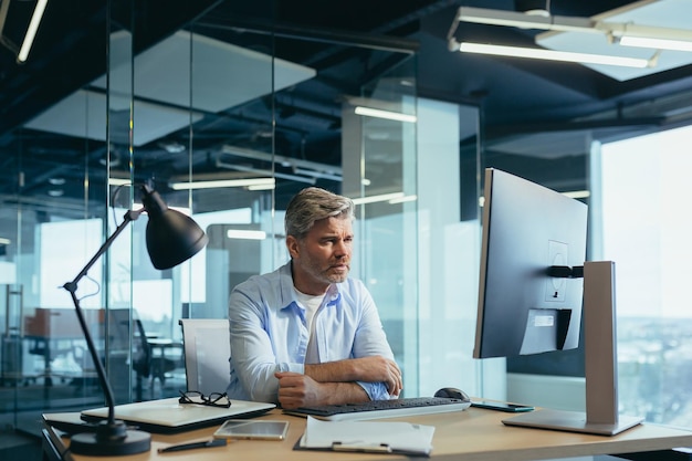 El hombre de negocios en pensamiento resuelve problemas importantes y toma decisiones. El hombre de negocios canoso trabaja en la computadora en la oficina moderna.