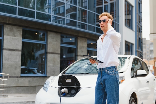 Hombre de negocios de pie cerca de cargar el coche eléctrico y usar la tableta en la calle.