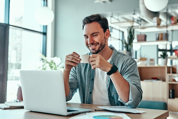 Hombre de negocios positivo mirando la pantalla del portátil en la cafetería Hombre sonriente bebiendo café en la cafetería Chico hablando de descanso entre el trabajo