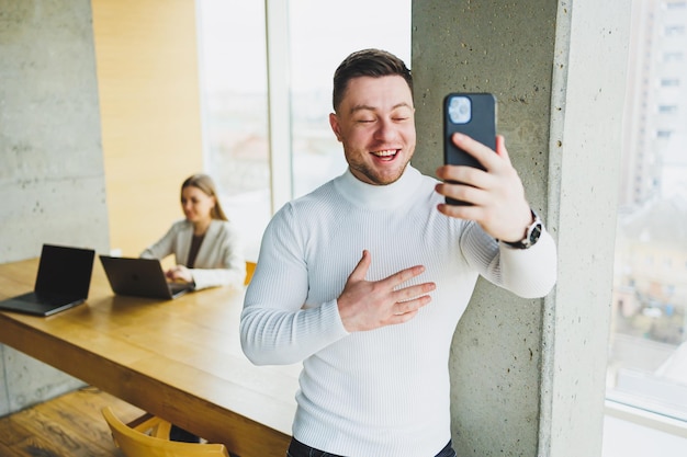 Hombre de negocios positivo en suéter sonriendo mirando el teléfono parado en una oficina moderna con teléfono trabajando en una empresa moderna