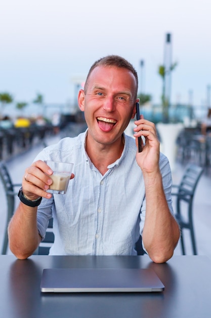 Hombre de negocios que trabaja con una computadora portátil en la terraza del hotel y habla por teléfono para administrar a los empleados durante las vacaciones en el resort Hombre en pantalones cortos y camisa bebiendo una bebida refrescante