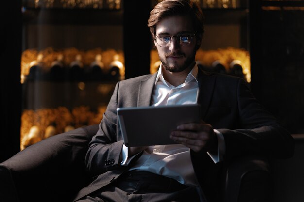 Hombre de negocios que trabaja con la tableta mientras está sentado en la cafetería a altas horas de la noche frente a las ventanas con vistas a la ciudad.