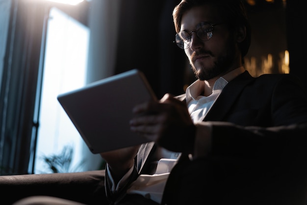 Hombre de negocios que trabaja con la tableta mientras está sentado en la cafetería a altas horas de la noche frente a las ventanas con vistas a la ciudad.