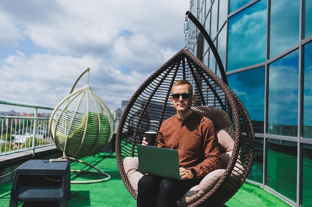 Un hombre de negocios con ropa formal se sienta en un sillón y mira una computadora portátil conectada a una red inalámbrica 4g en la terraza de una oficina moderna mientras trabaja de forma remota