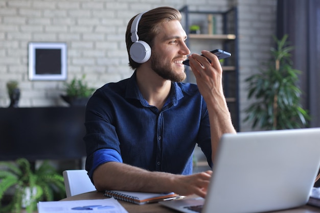 Hombre de negocios satisfecho con gafas sosteniendo un teléfono inteligente cerca de la boca para grabar mensajes de voz o chatear en línea.