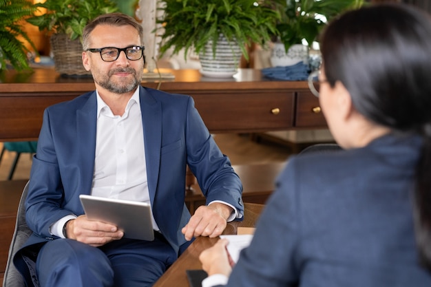 Hombre de negocios seguro bien vestido con tableta sentado junto a la mesa en el restaurante mientras interactúa con colegas en la reunión