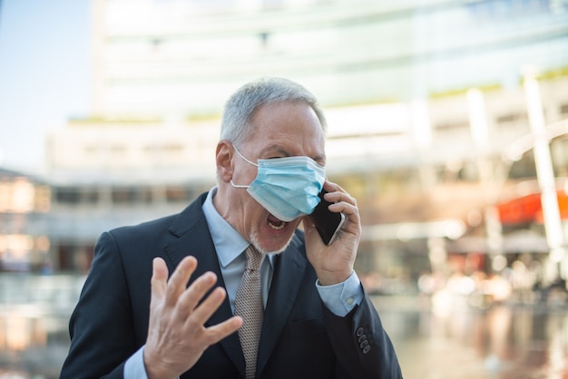 Hombre de negocios senior enmascarado enojado gritando al teléfono al aire libre, covid y concepto de coronavirus