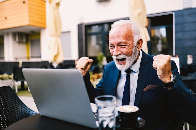 Un hombre de negocios senior se sienta en un café mirando su computadora portátil y celebrando el éxito