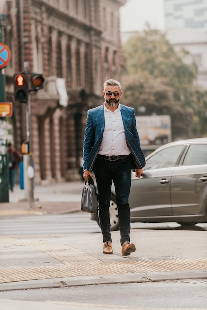 Un hombre de negocios senior con traje y gafas de sol caminando por la ciudad mientras lleva una bolsa