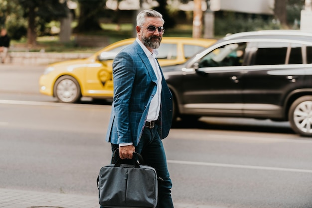 Un hombre de negocios senior con traje y gafas de sol caminando por la ciudad mientras lleva una bolsa