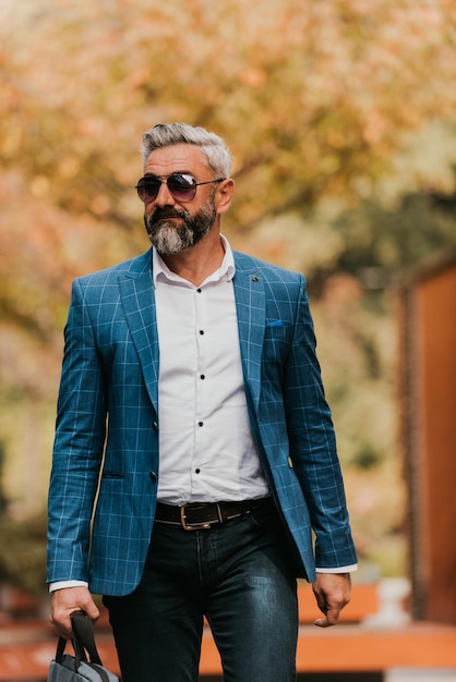 Un hombre de negocios senior con traje y gafas de sol caminando por la ciudad mientras lleva una bolsa