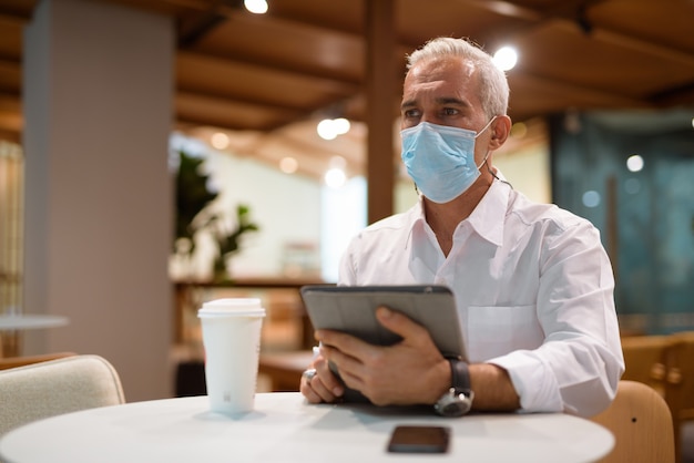 Hombre de negocios sentado en la cafetería y usando tableta digital mientras usa mascarilla y tiro horizontal de distanciamiento social