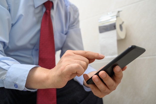 Hombre de negocios sentado en el inodoro en el baño y usando un teléfono inteligente.