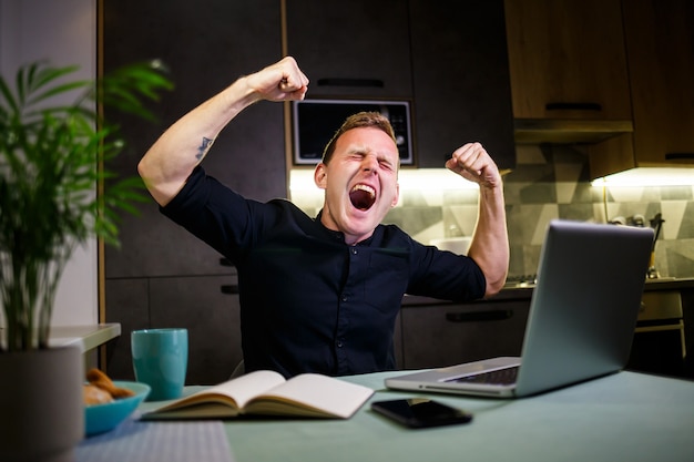 Hombre de negocios sentado en la mesa de casa, casa moderna y acogedora, mirando la pantalla del portátil, se siente satisfecho con el orgullo por el trabajo realizado, el hombre exitoso se regocija, levantando las manos detrás de la cabeza