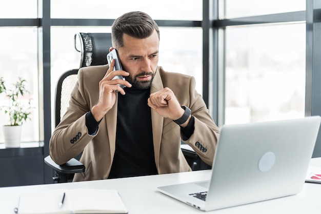 Un hombre de negocios serio está hablando por su teléfono celular y mirando su computadora portátil