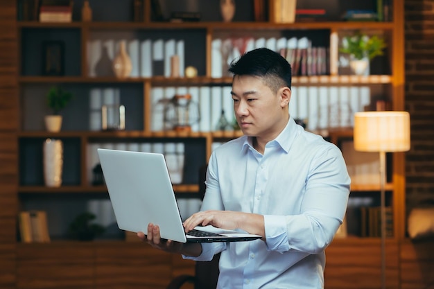 Un hombre de negocios serio de pie trabajando en una laptop asiática mirando la pantalla enfocada en leer e investigar datos económicos