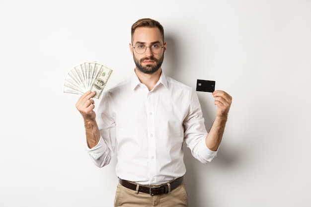 Hombre de negocios serio que mira la cámara, sosteniendo la tarjeta de crédito y el dinero, de pie sobre el fondo blanco. Concepto de compras y finanzas.