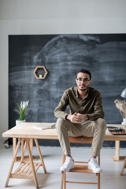 Hombre de negocios serio en ropa casual y anteojos mirándote mientras está sentado en la mesa con las piernas en la silla de madera