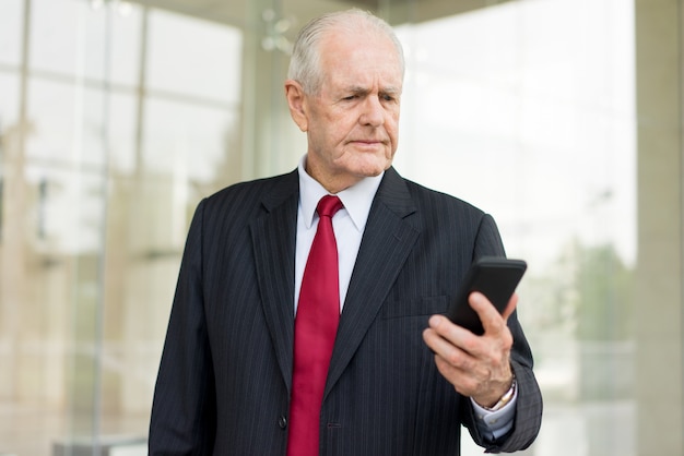hombre de negocios serio senior leyendo un mensaje en el teléfono inteligente con oficina borrosa