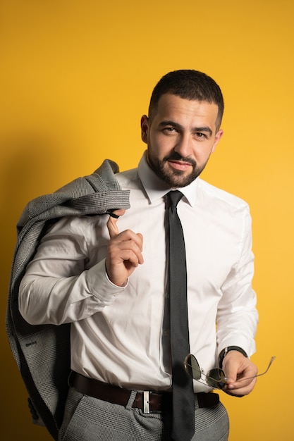 Hombre de negocios serio vestido con traje gris posando sosteniendo su chaqueta en el hombro colgando detrás y gafas de sol en la otra mano mirando al frente con una sonrisa aislada en la pared amarilla