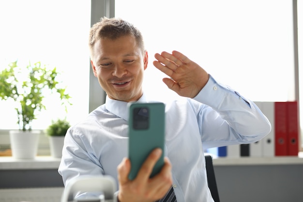 El hombre de negocios se sienta en el lugar de trabajo y saluda con la mano al teléfono inteligente. Comunicación empresarial en línea por concepto de videollamada