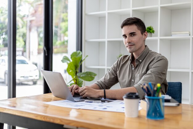 Foto un hombre de negocios se sienta en su oficina con una computadora portátil y material de oficina, en su escritorio hay documentos financieros que está revisando, está escribiendo, enviando mensajes de texto, hablando con su socio a través de un mensajero de computadora portátil.