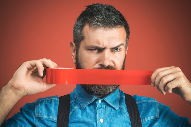 Hombre de negocios silenciado con cinta adhesiva sobre su boca. Hombre barbudo con cinta adhesiva alrededor de la boca. Infeliz hombre en camisa de mezclilla cubrió la boca con cinta aislante roja. Aislado sobre fondo rojo.