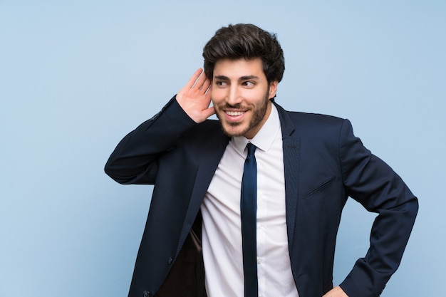 Hombre de negocios sobre la pared azul aislada que escucha algo poniendo la mano en la oreja