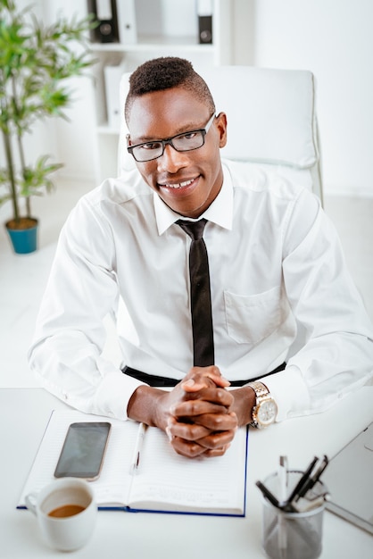 Hombre de negocios sonriente africano con gafas sentado en la oficina y mirando a la cámara.